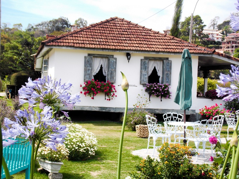 foto-apresenta-uma-bela-casinha-com-jardineiras-penduradas-na-janela-e-o-colorido-da-flores-dão-um-ar-de-clima-europeu
